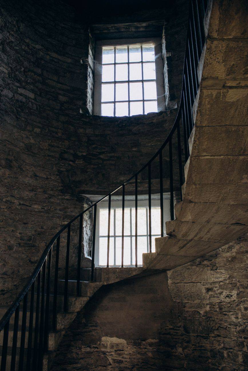 A dimly lit stone spiral staircase with metal railings and barred windows creates a mysterious atmosphere.