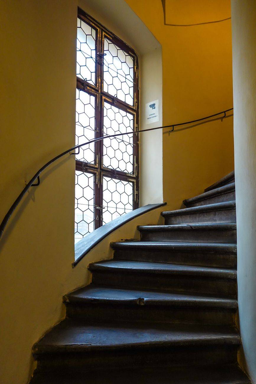 A curved staircase leading upwards, next to a vintage window with hexagonal patterns in a historic building.