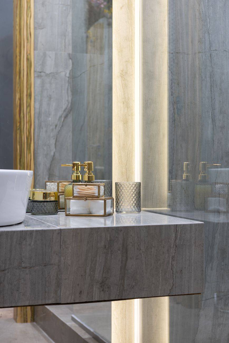 Stylish washroom with gray tiled walls and different perfume placed on marble counter near sink with reflective mirror at home