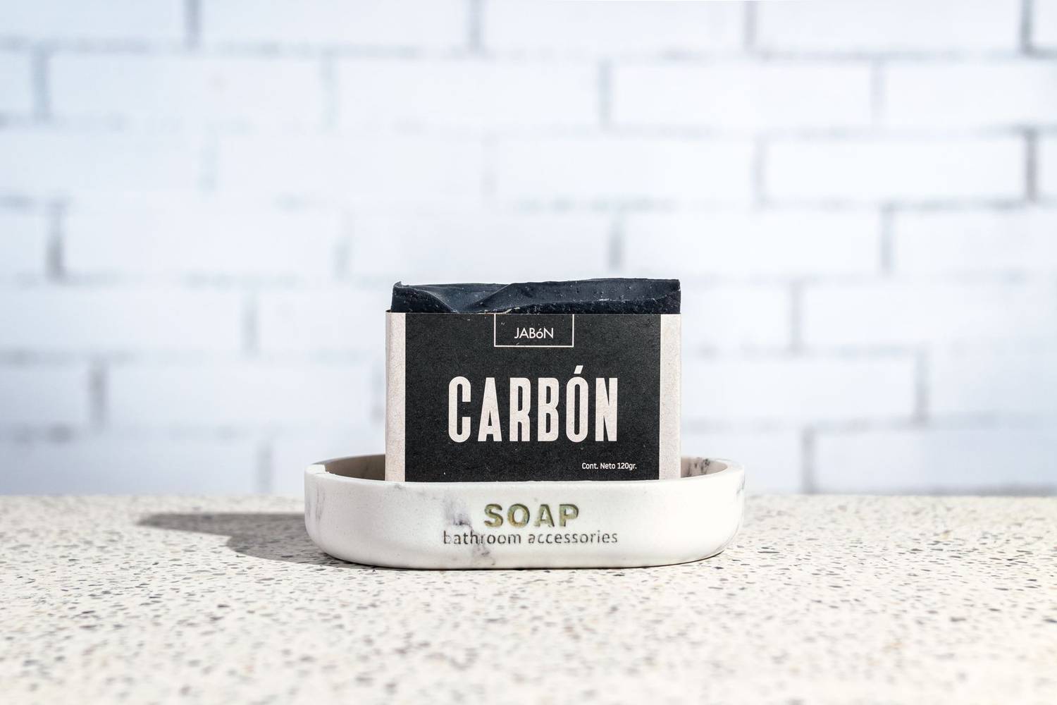 Close-up of handcrafted charcoal soap on a marble tray against a white brick wall.