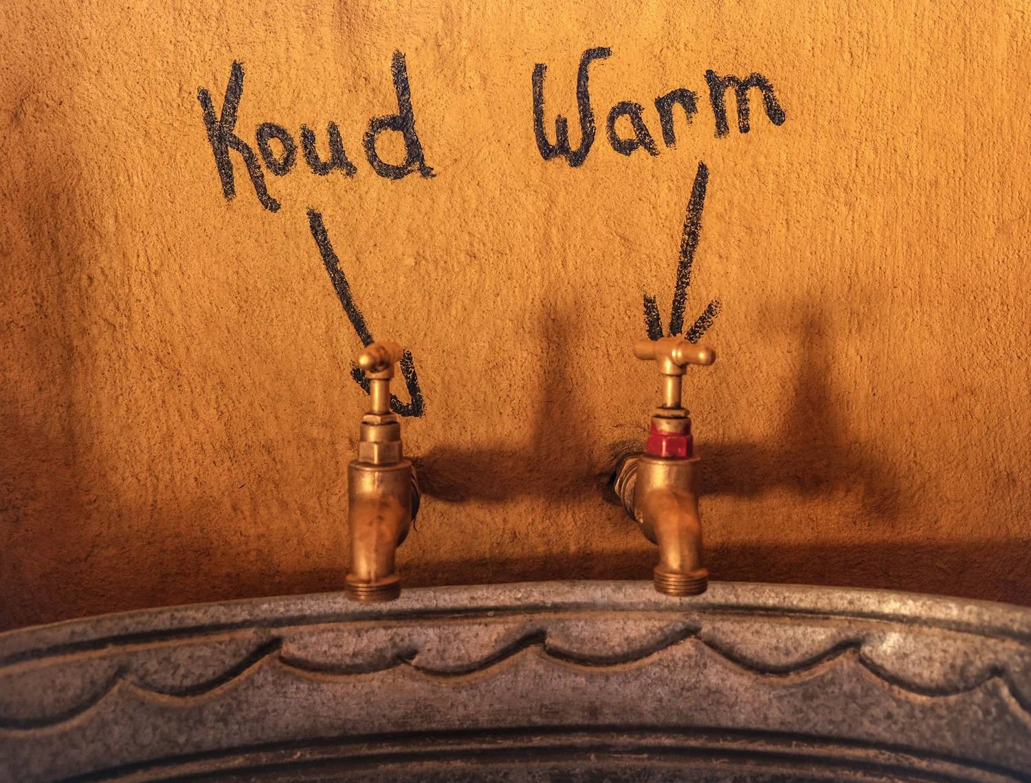 Close-up of vintage metal faucets labeled cold 'Koud' and warm 'Warm' on a textured gold wall.
