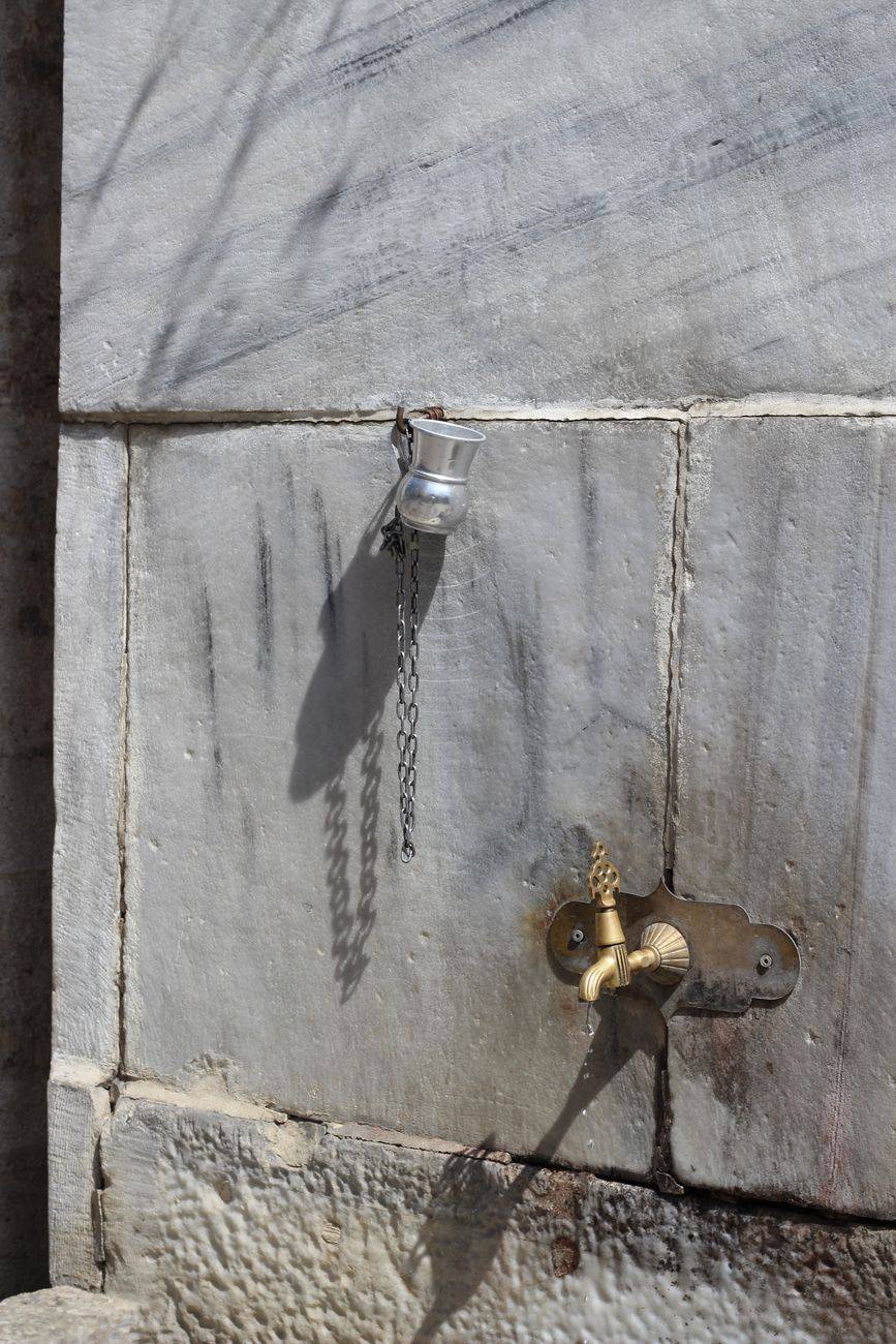 A classic outdoor fountain with a metal cup and brass tap on a stone wall.