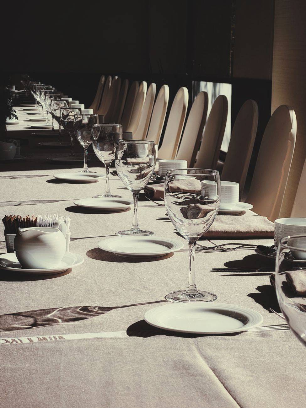 Long dining table elegantly set with glassware and plates in a sunlit room.
