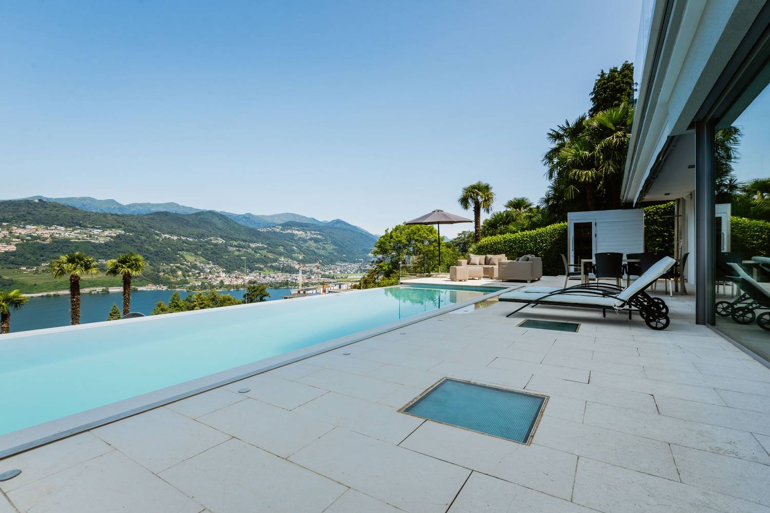 Infinity pool with loungers and umbrella overlooking lake and mountains on a sunny day.