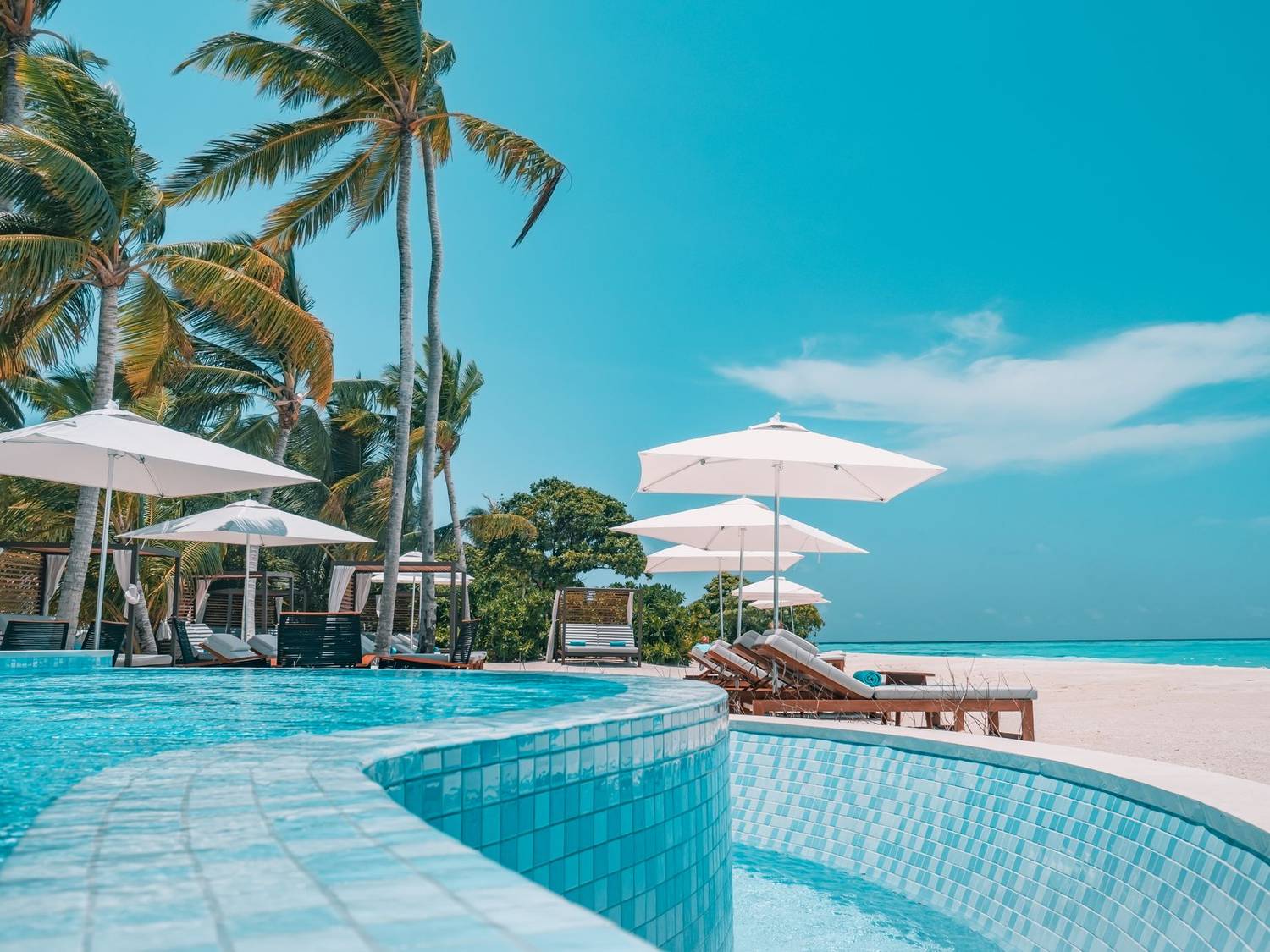 Tiled pool of exotic resort placed on white sandy beach with parasols and green palms