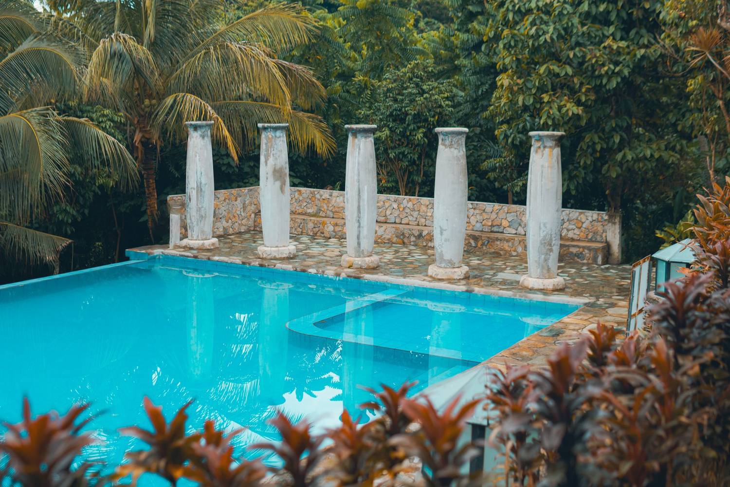 A tranquil tropical poolside with ancient columns surrounded by lush greenery.