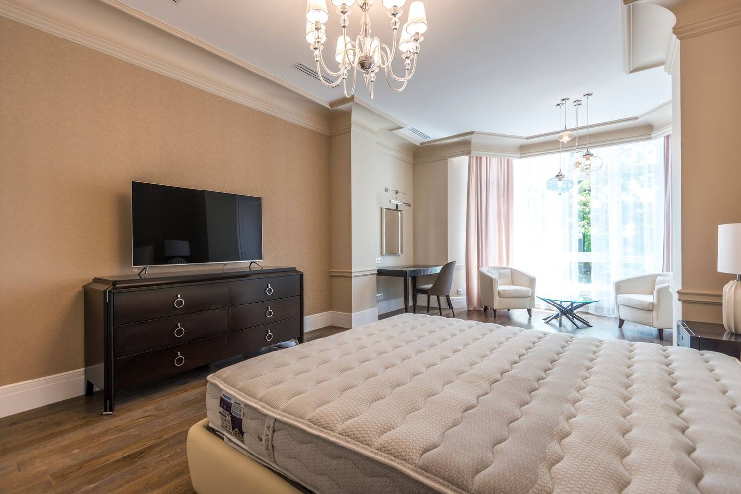 Spacious modern bedroom with chandelier and flat screen TV, featuring a minimalist and elegant design.