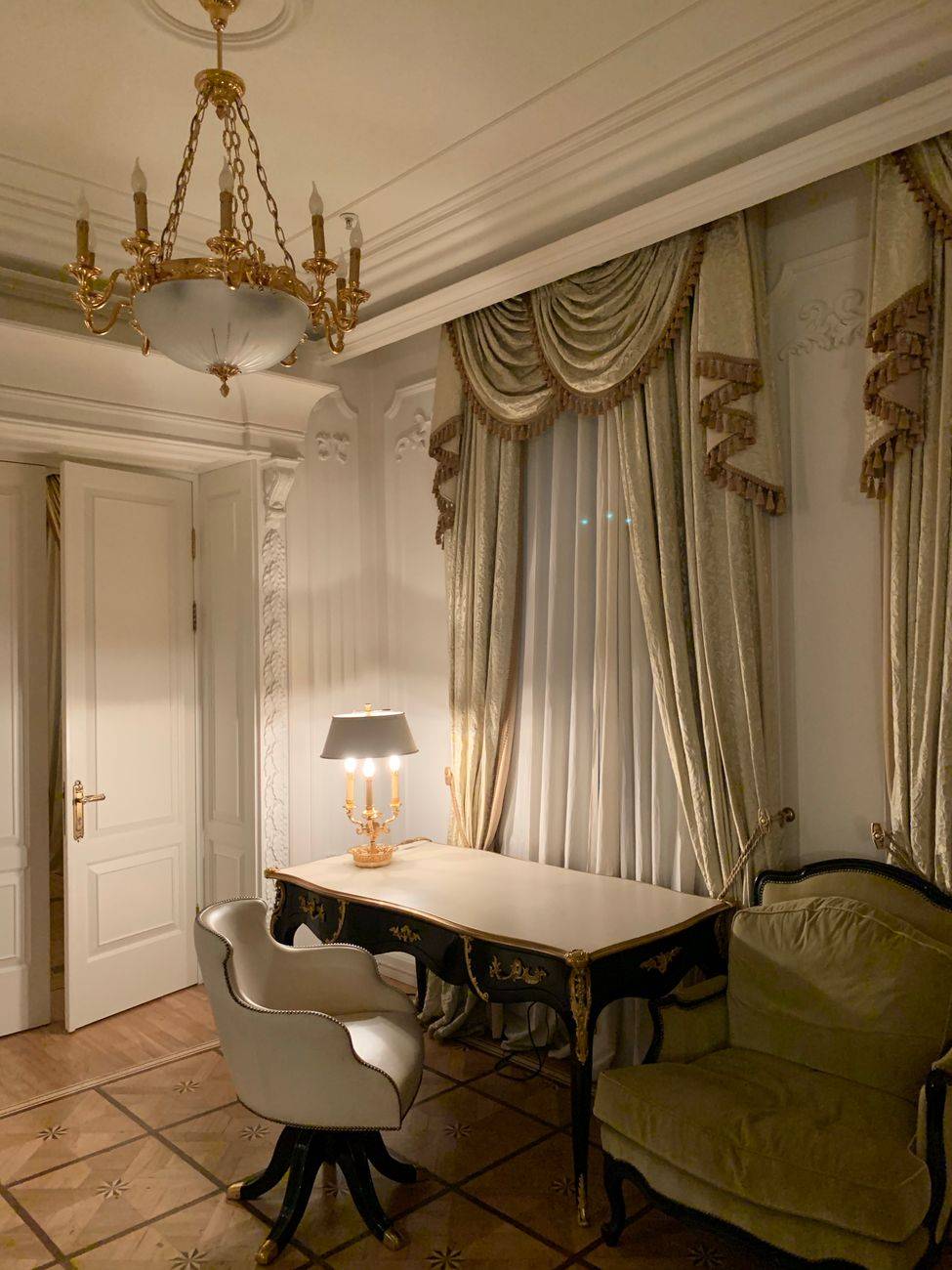 Luxurious hotel room interior featuring a classic chandelier, ornate curtains, and a vintage desk.