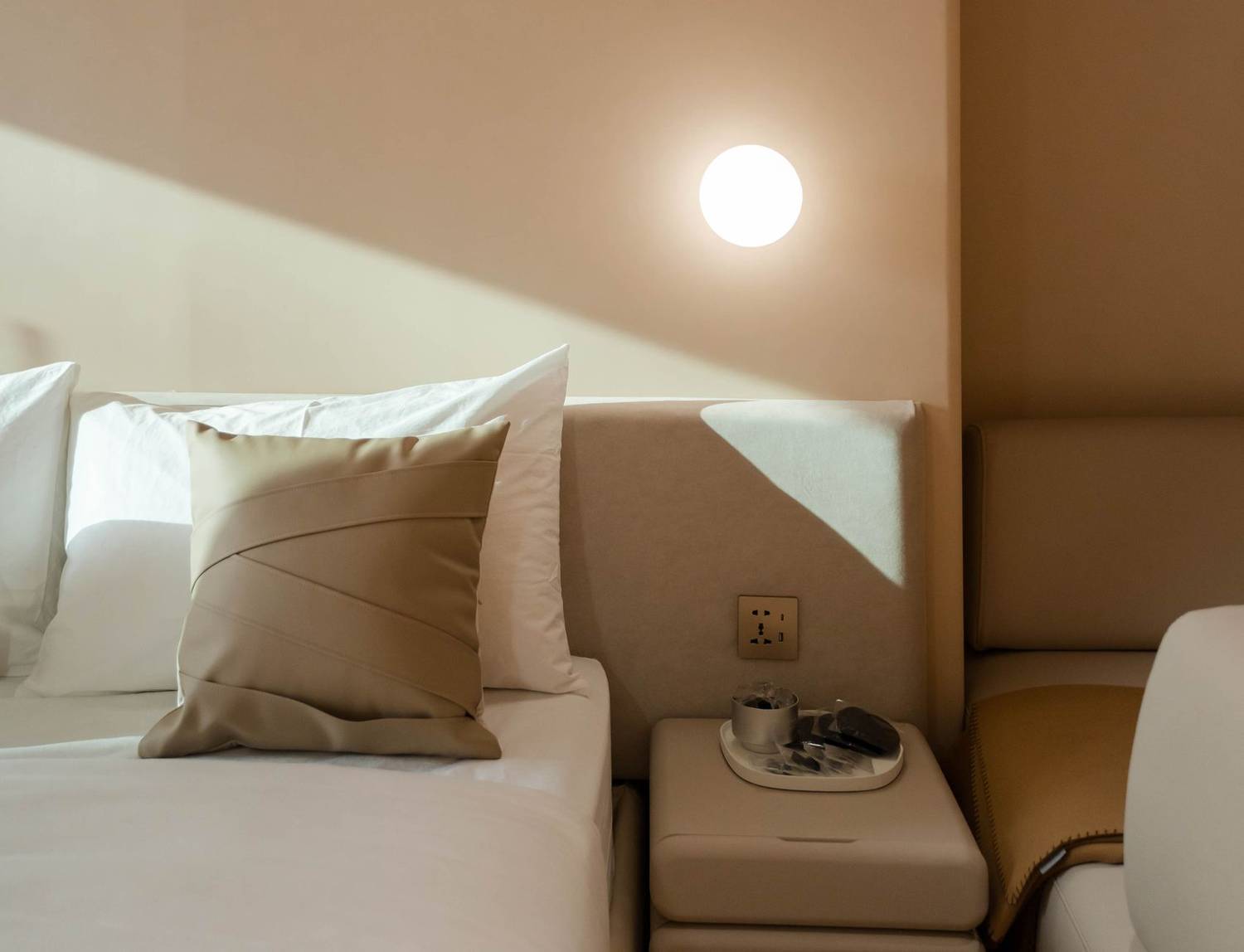 A modern bedroom with beige tones, featuring a cushioned bed, elegant pillows, and soft wall lighting.