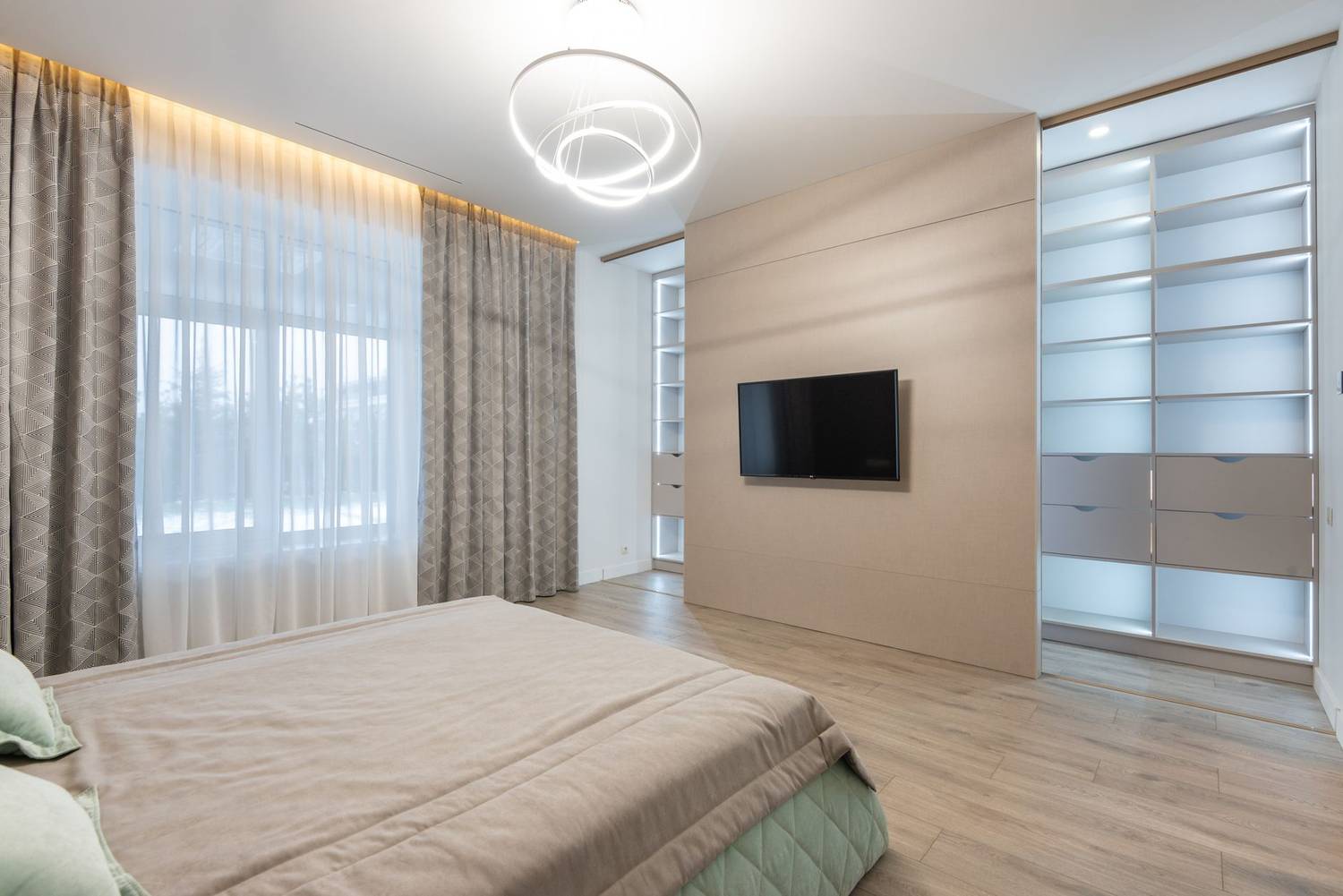 Inside bedroom interior furnished with comfortable bed covered with soft plaid against TV set placed on wall near big wardrobe with glass pa