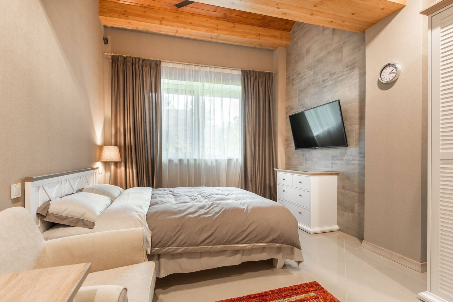 Interior of modern hotel room with comfortable bed near window with curtains and white sofa against tv and dresser