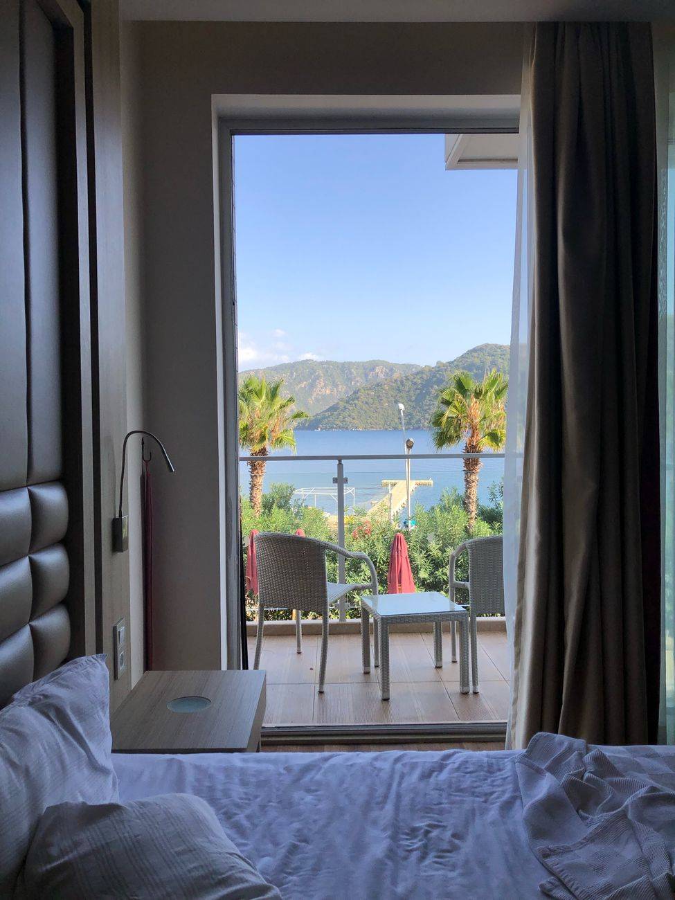 Cozy bedroom with balcony view of palm trees, a lake, and mountains in the distance.