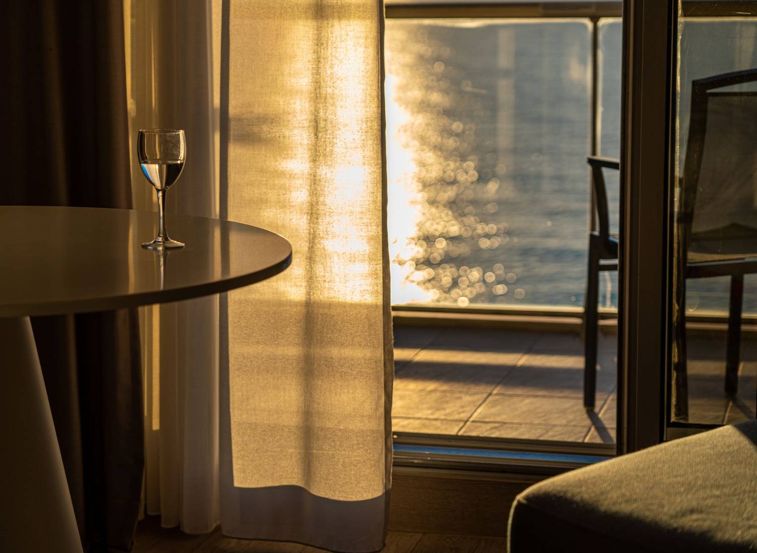 Sunlit room with a wine glass on table, overlooking a serene body of water from a balcony.
