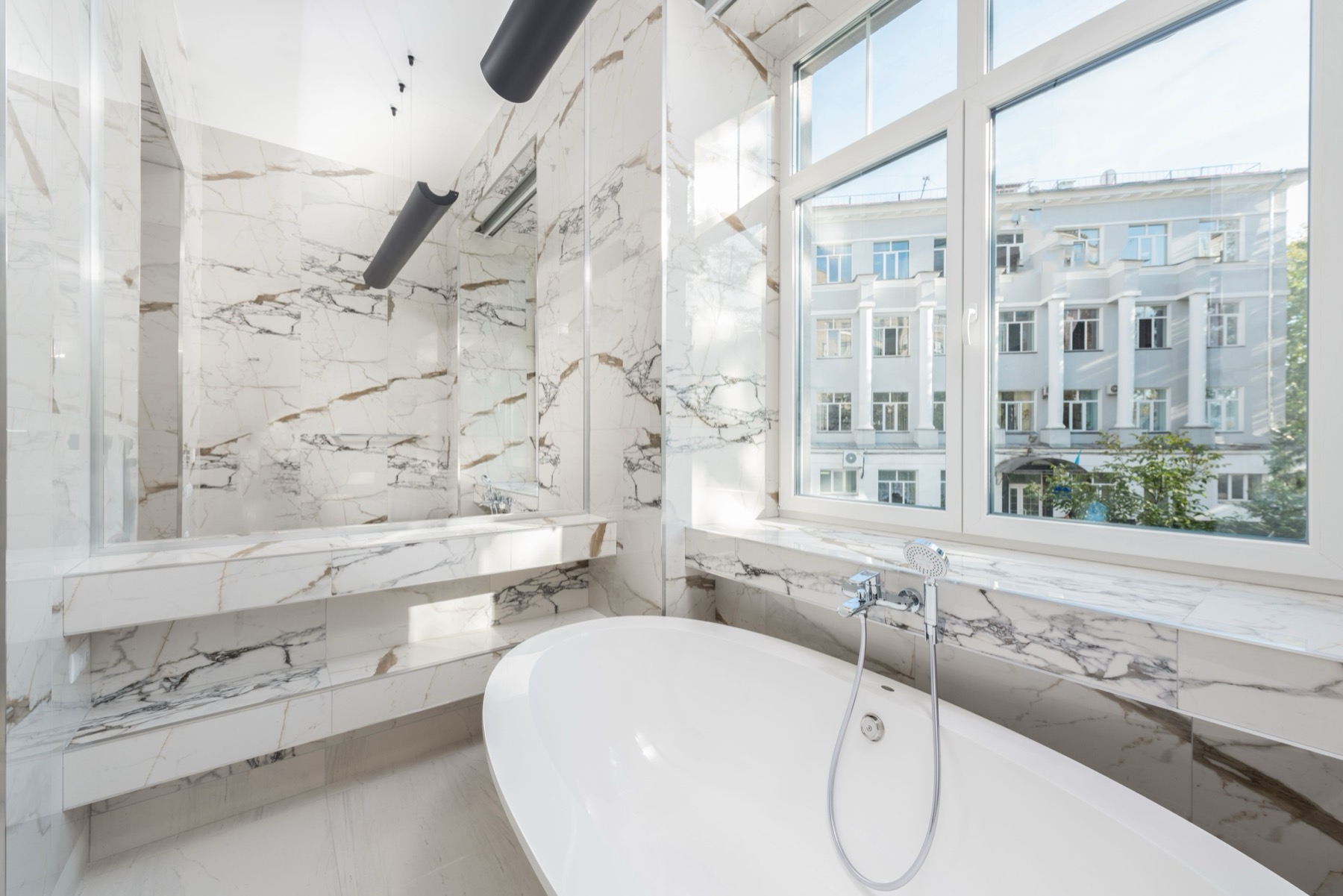 A pale Calacatta marble bathroom with a freestanding tub beside a tall window overlooking a Haussmann facade
